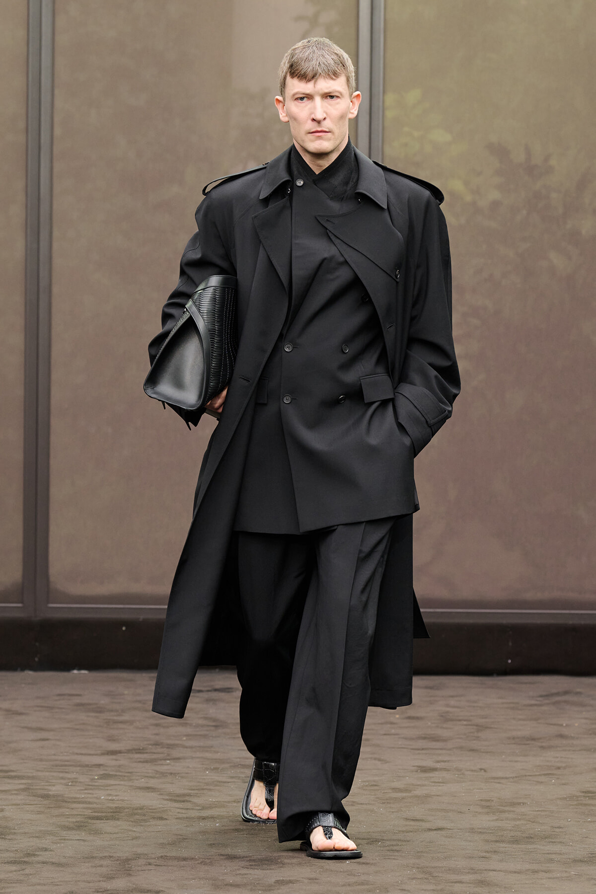 Man walking outside in an all-black, layered outfit: long coat over a double-breasted suit, carrying a black textured portfolio under his arm.