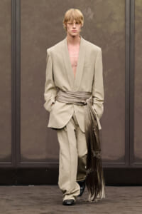Model on runway wearing an oversized beige suit with a deep V-neck and a tan sash tied at the waist.