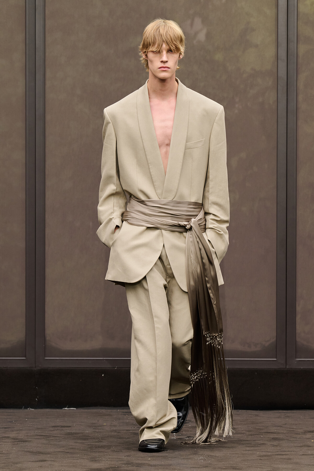 Model on runway wearing an oversized beige suit with a deep V-neck and a tan sash tied at the waist.
