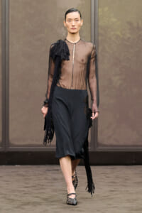Model walks a runway wearing a sheer black blouse with fringe on the left shoulder and black wide-leg midi pants, pairing with strappy heels.