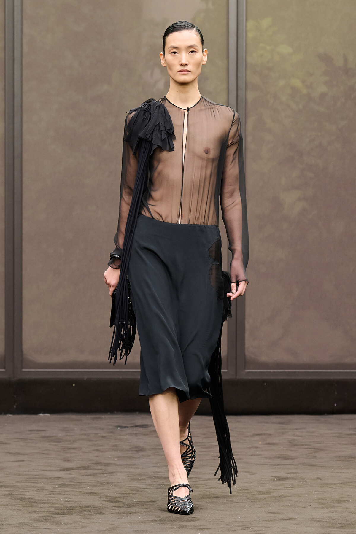 Model walks a runway wearing a sheer black blouse with fringe on the left shoulder and black wide-leg midi pants, pairing with strappy heels.