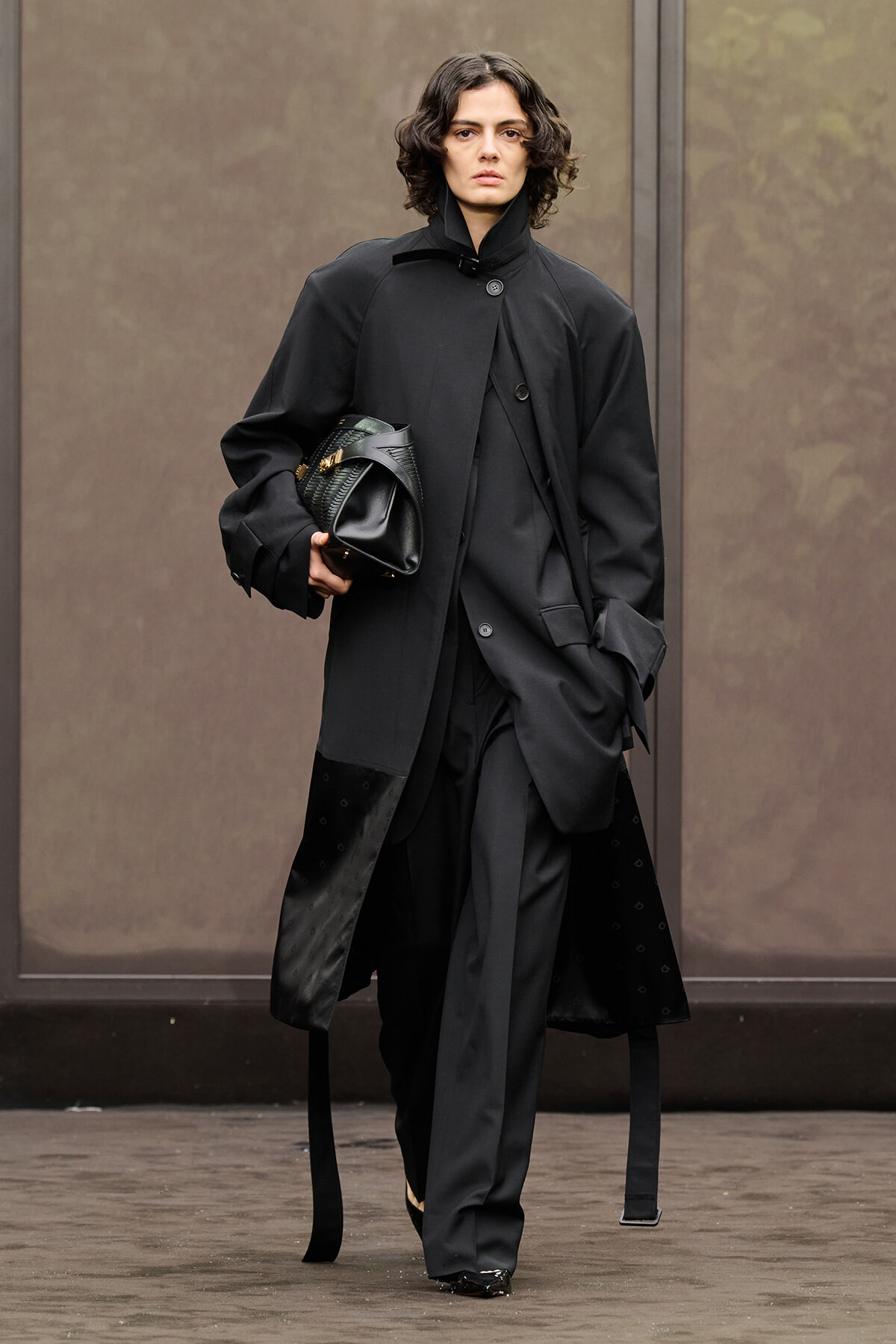 Model in an all-black, oversized coat walking outdoors while holding a black handbag with gold accents.