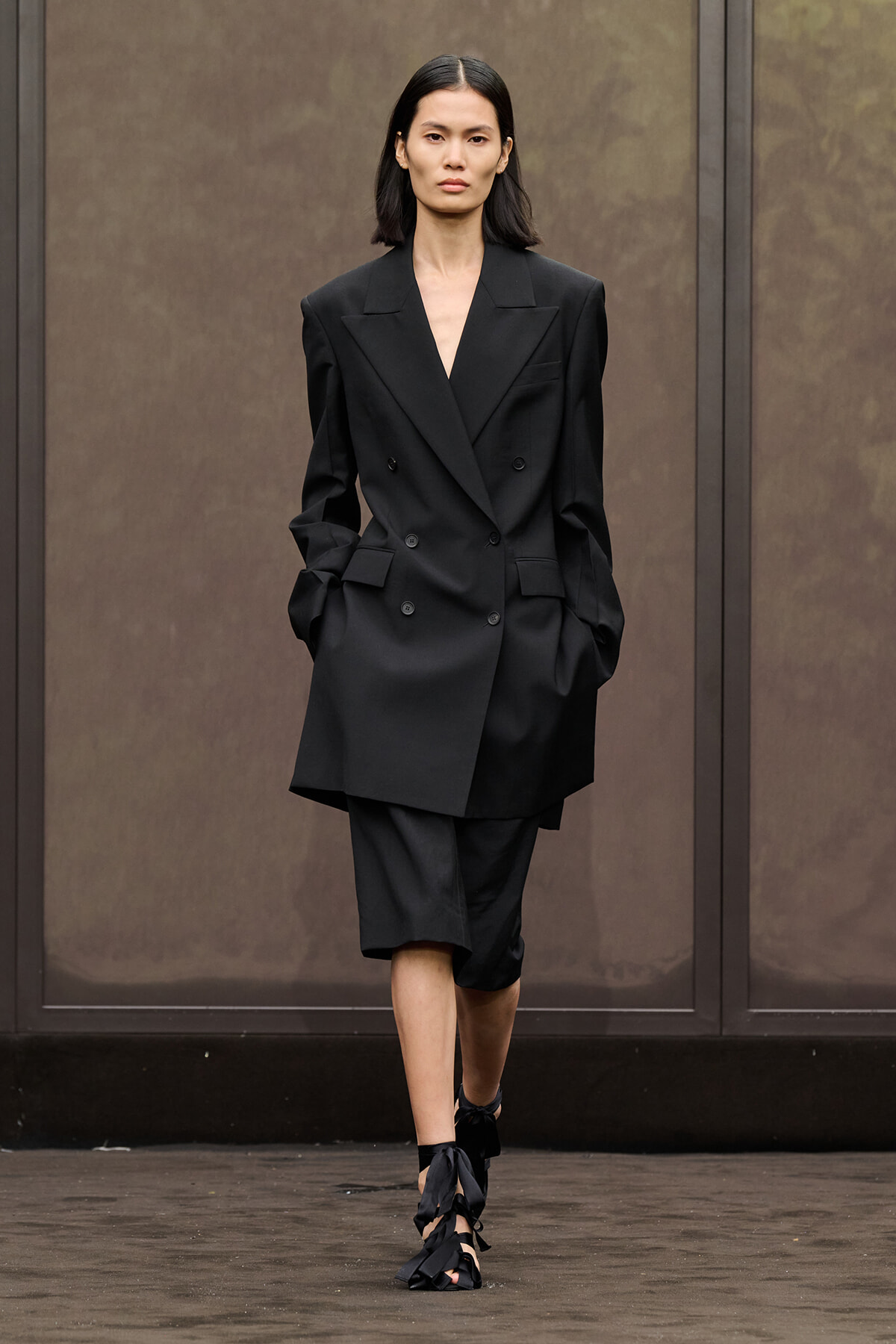 Model walking in a long black double-breasted blazer and matching knee-length shorts, black strappy heeled sandals, neutral expression.