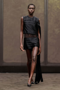Model on a runway wearing a black, asymmetrical dress with layered panels and a sheer lace waist, paired with metallic pointed heels.