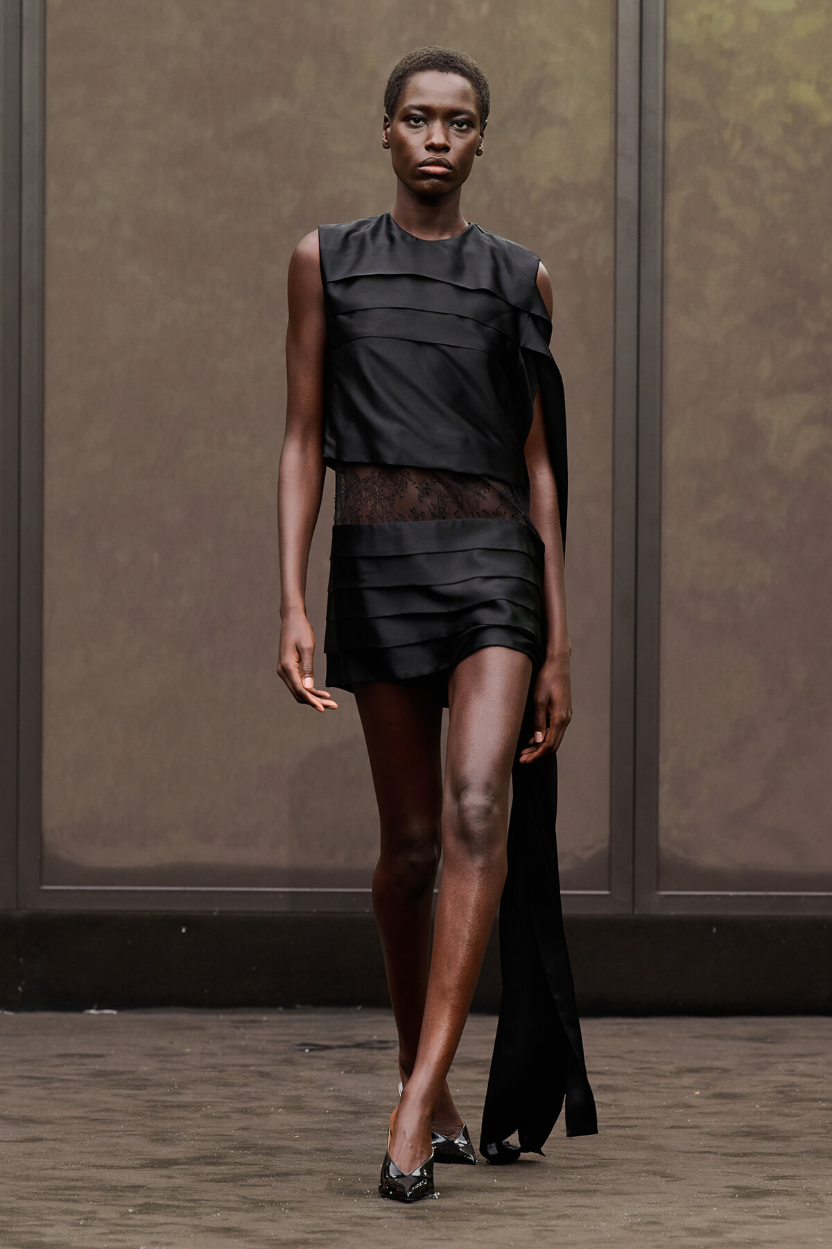 Model on a runway wearing a black, asymmetrical dress with layered panels and a sheer lace waist, paired with metallic pointed heels.
