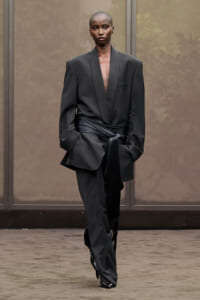 Model walks the runway in an oversized black blazer with a deep V neckline and a wrapped waist sash over matching pants.