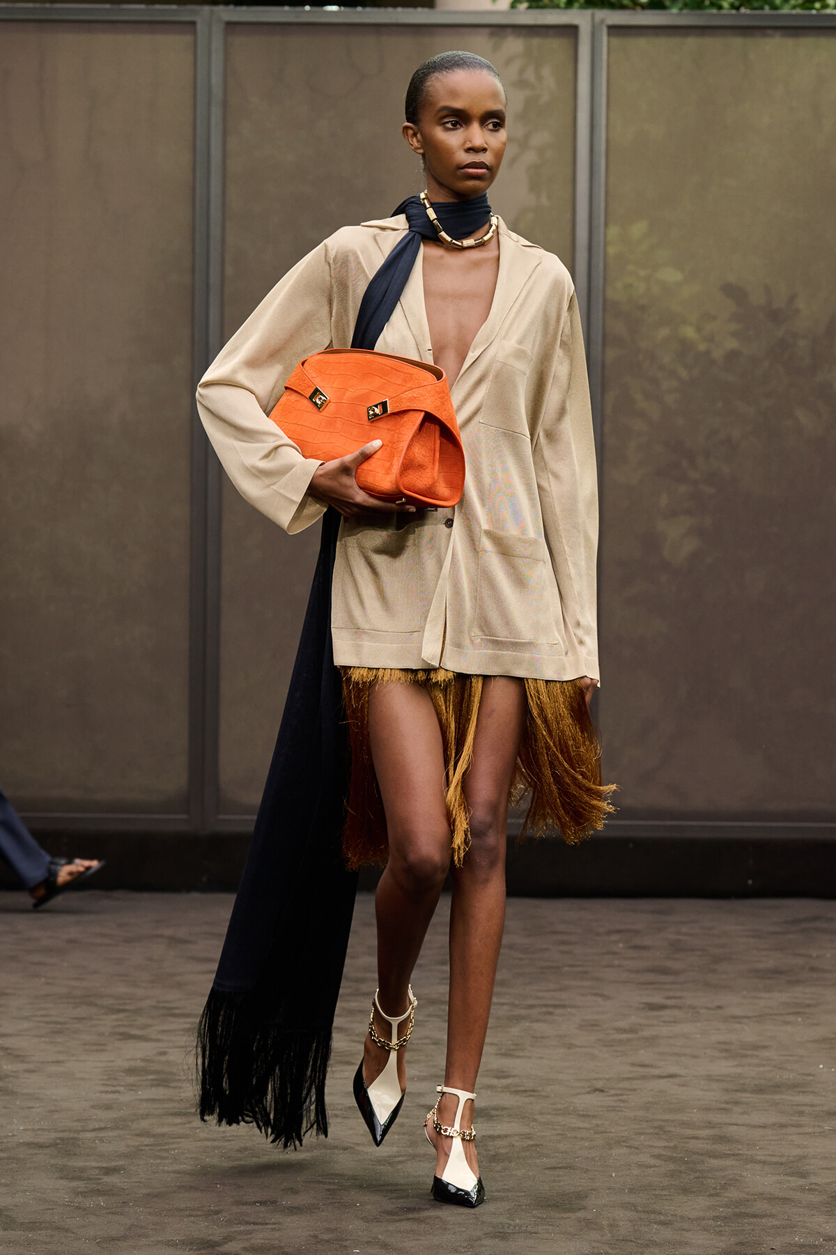 Model on the runway wearing a tan oversized blazer, black scarf, orange handbag, and a fringe hem skirt with high heels.