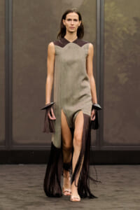 Model walks a runway in a taupe-grey sleeveless dress with a brown scalloped neckline and long black fringe accessories, wearing nude sandals.