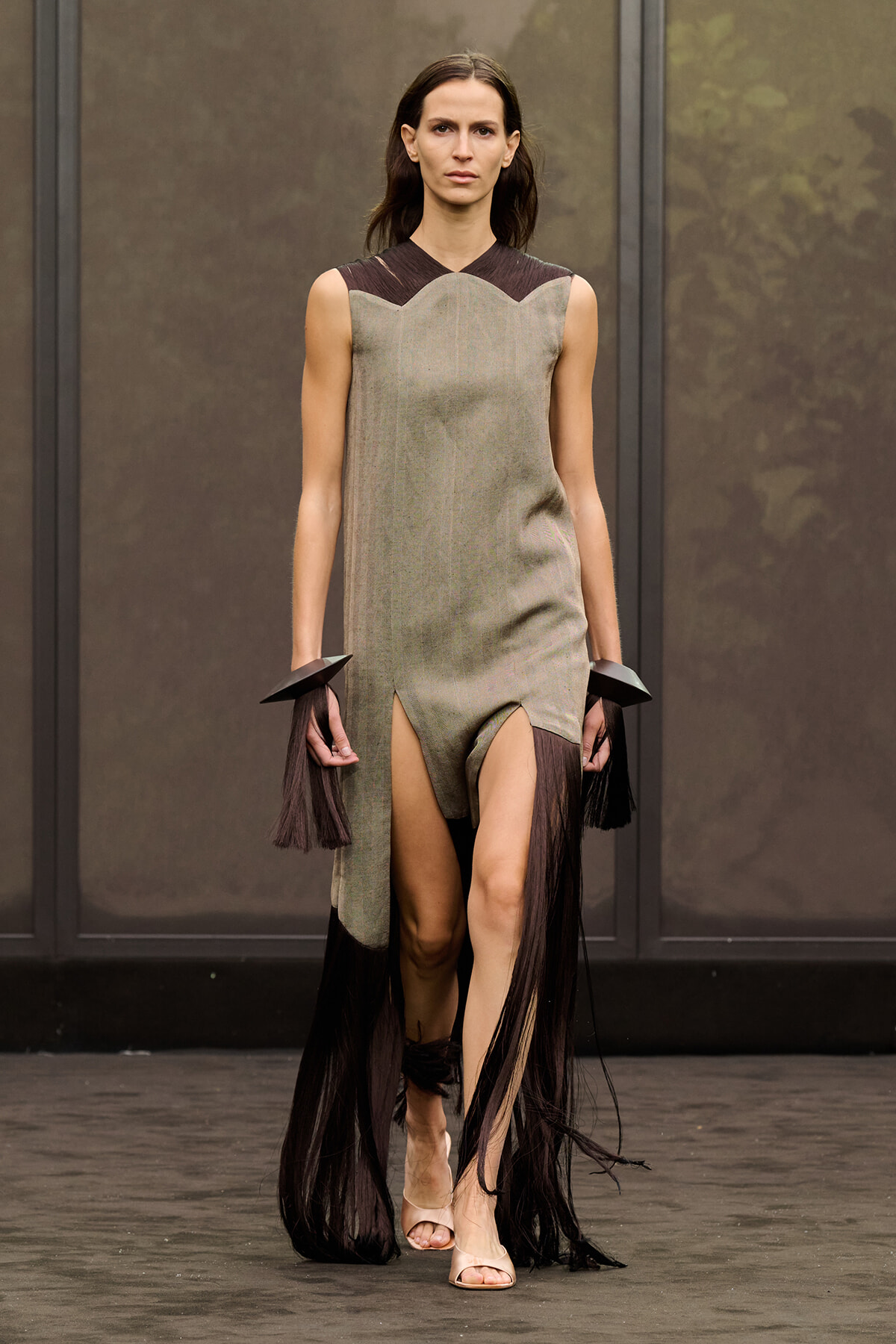 Model walks a runway in a taupe-grey sleeveless dress with a brown scalloped neckline and long black fringe accessories, wearing nude sandals.