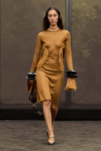 Model walks the runway in a camel-colored dress with fringe panels at the sides and long sleeves, paired with bold black cuff accessories and heels, carrying a bag.