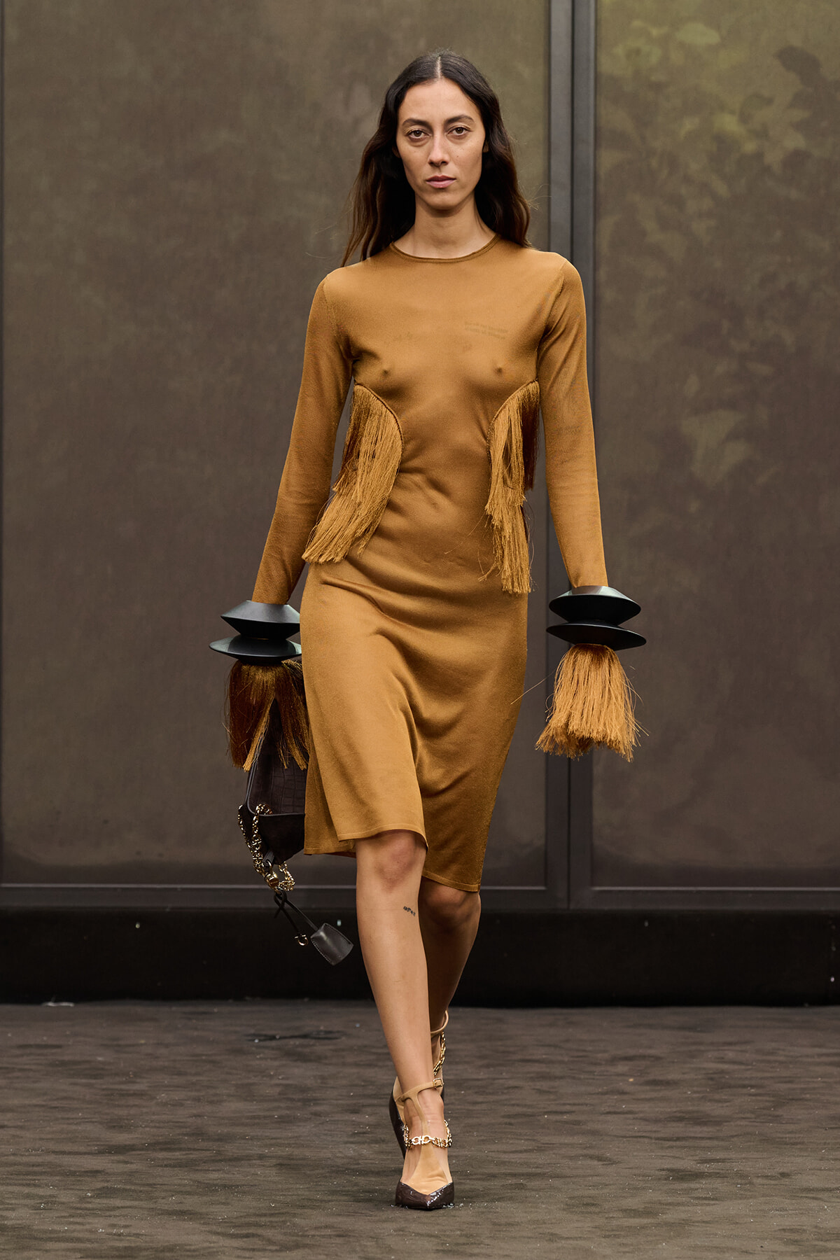 Model walks the runway in a camel-colored dress with fringe panels at the sides and long sleeves, paired with bold black cuff accessories and heels, carrying a bag.