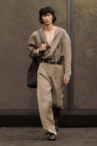 Male model walking in a beige utility jumpsuit with a brown belt, carrying a dark brown tote over the shoulder.