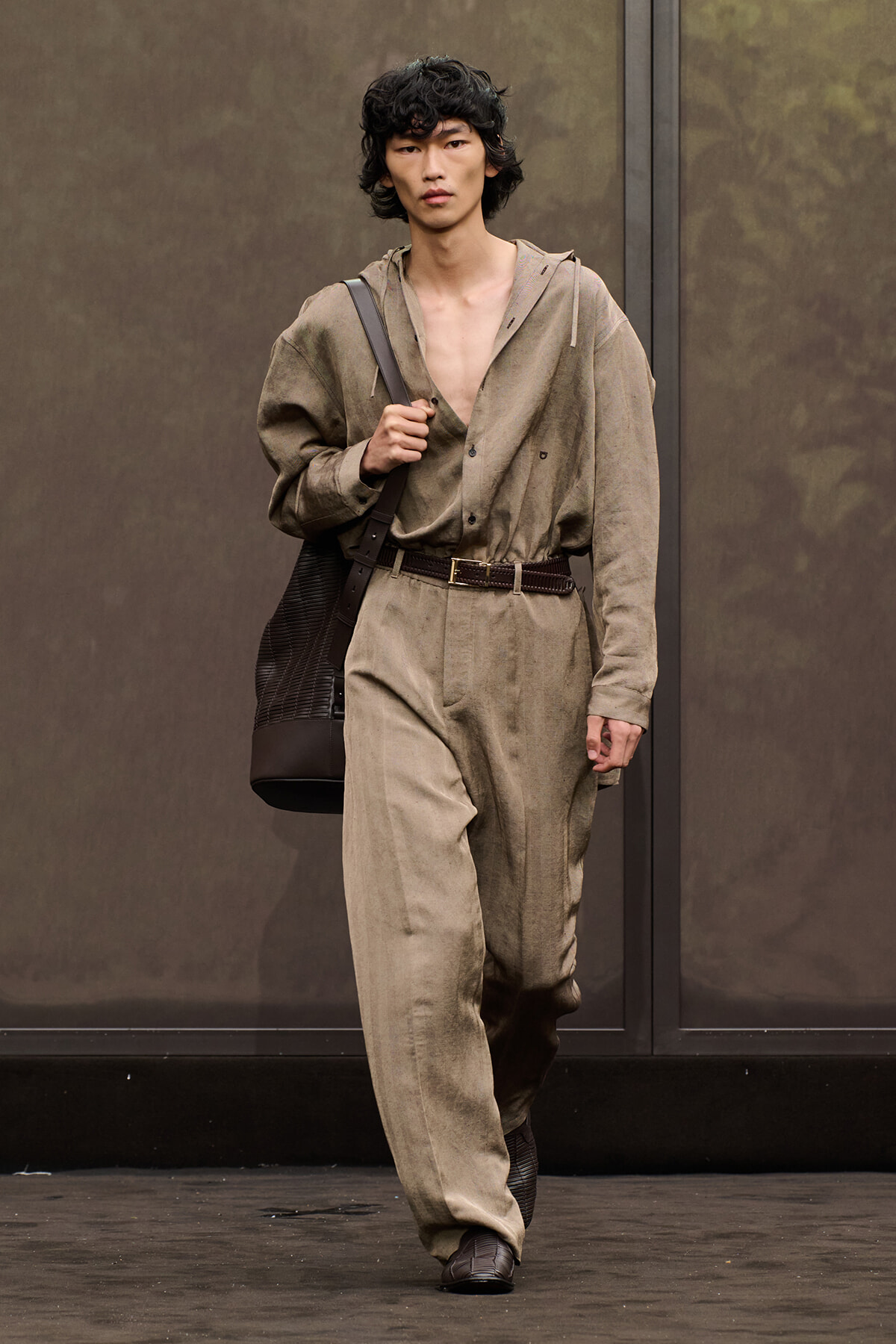 Male model walking in a beige utility jumpsuit with a brown belt, carrying a dark brown tote over the shoulder.