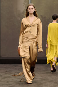 Model walks the runway in a gold satin suit with a wrapped belt and matching pants, carrying a small brown bag.