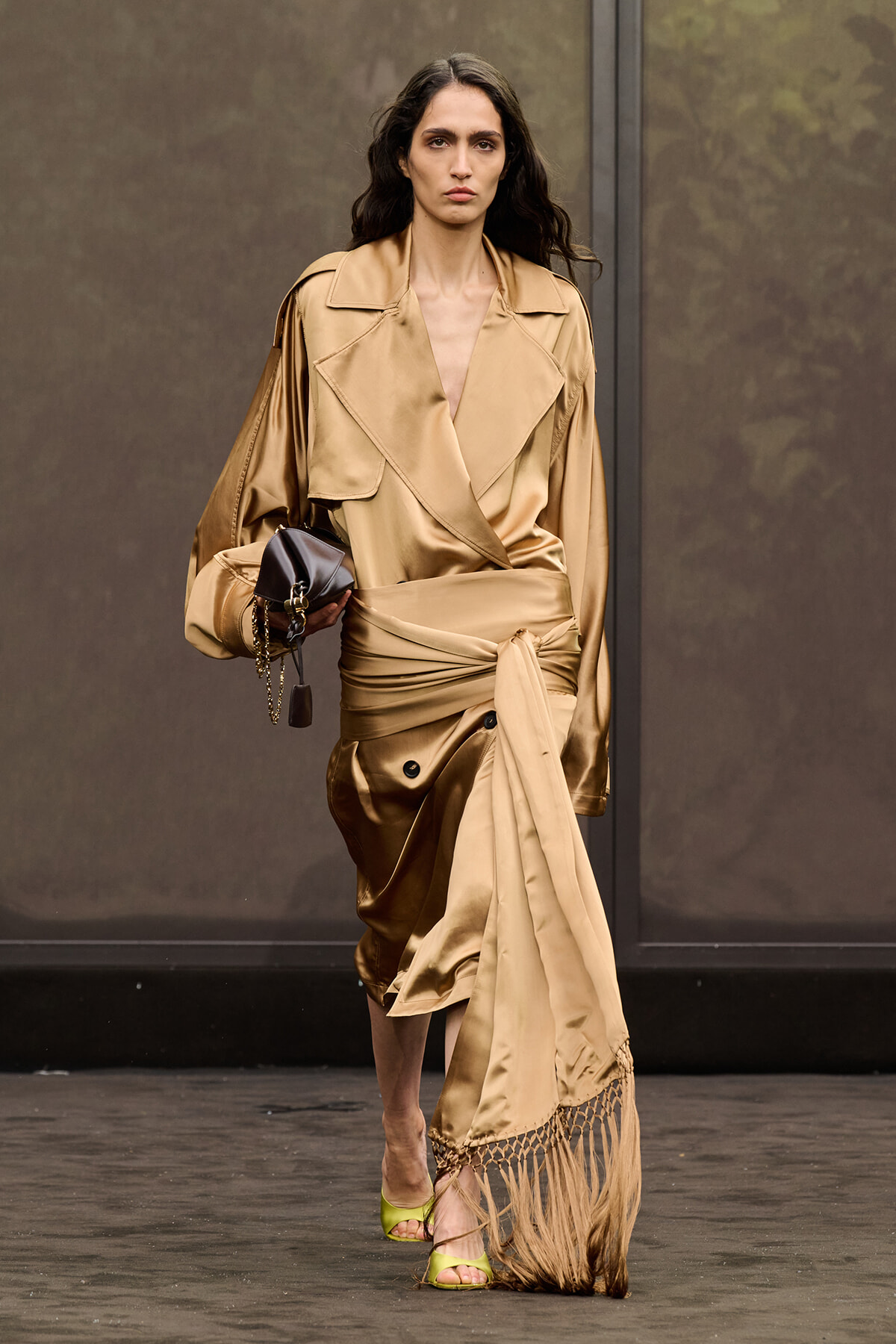 Model walking the runway in a gold satin wrap dress with fringe hem and a small black handbag.