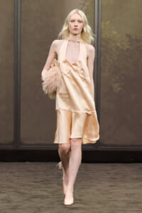 Model walking a runway in a peach satin halter dress with a keyhole neckline, carrying a fluffy feathered clutch.