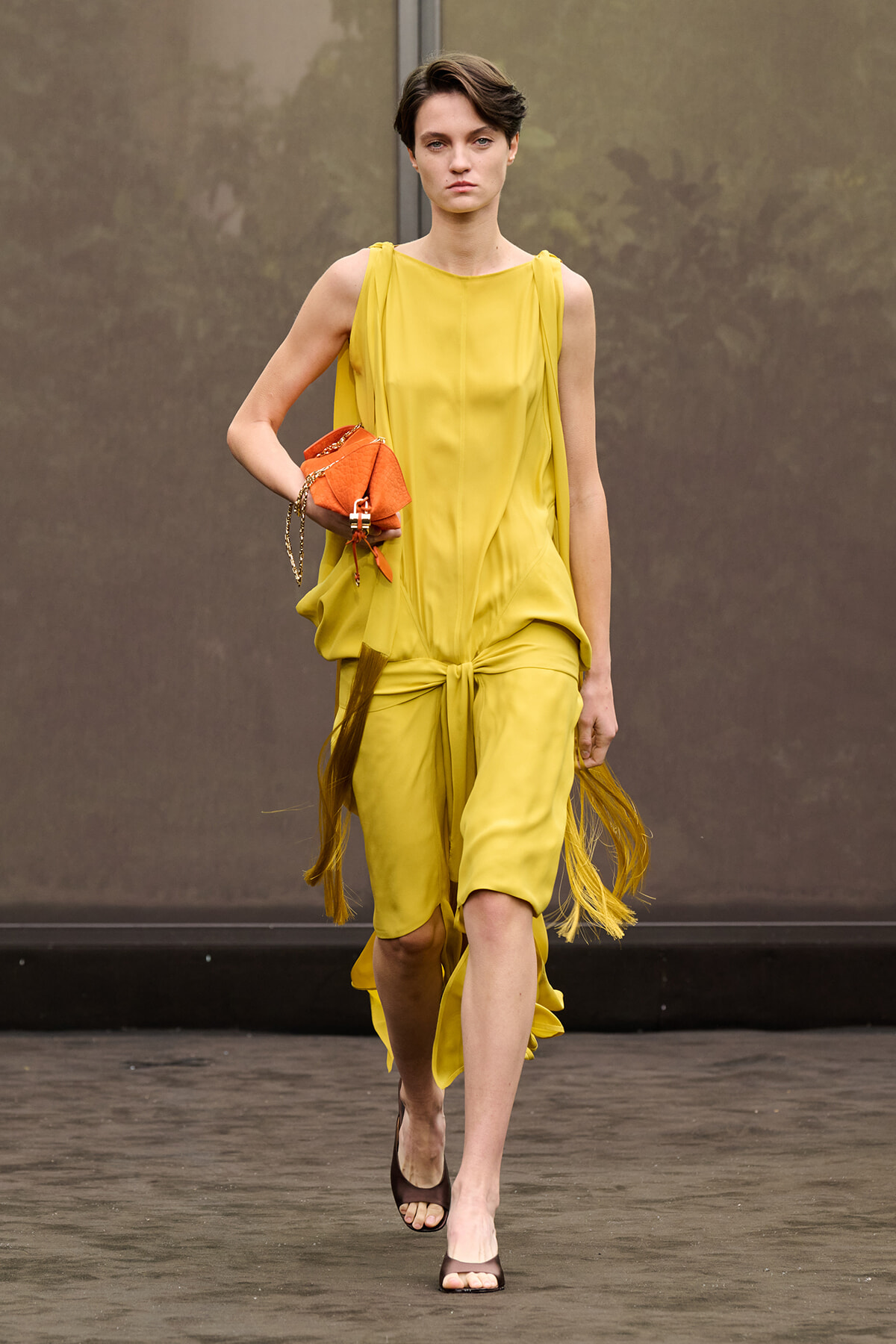 Female model walking a runway in a bright yellow sleeveless dress with a knot at the waist, holding an orange handbag and wearing brown sandals.