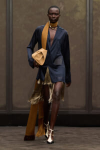 Model struts in a navy blazer-dress with gold fringe, tan scarf, and beige bag in hand.