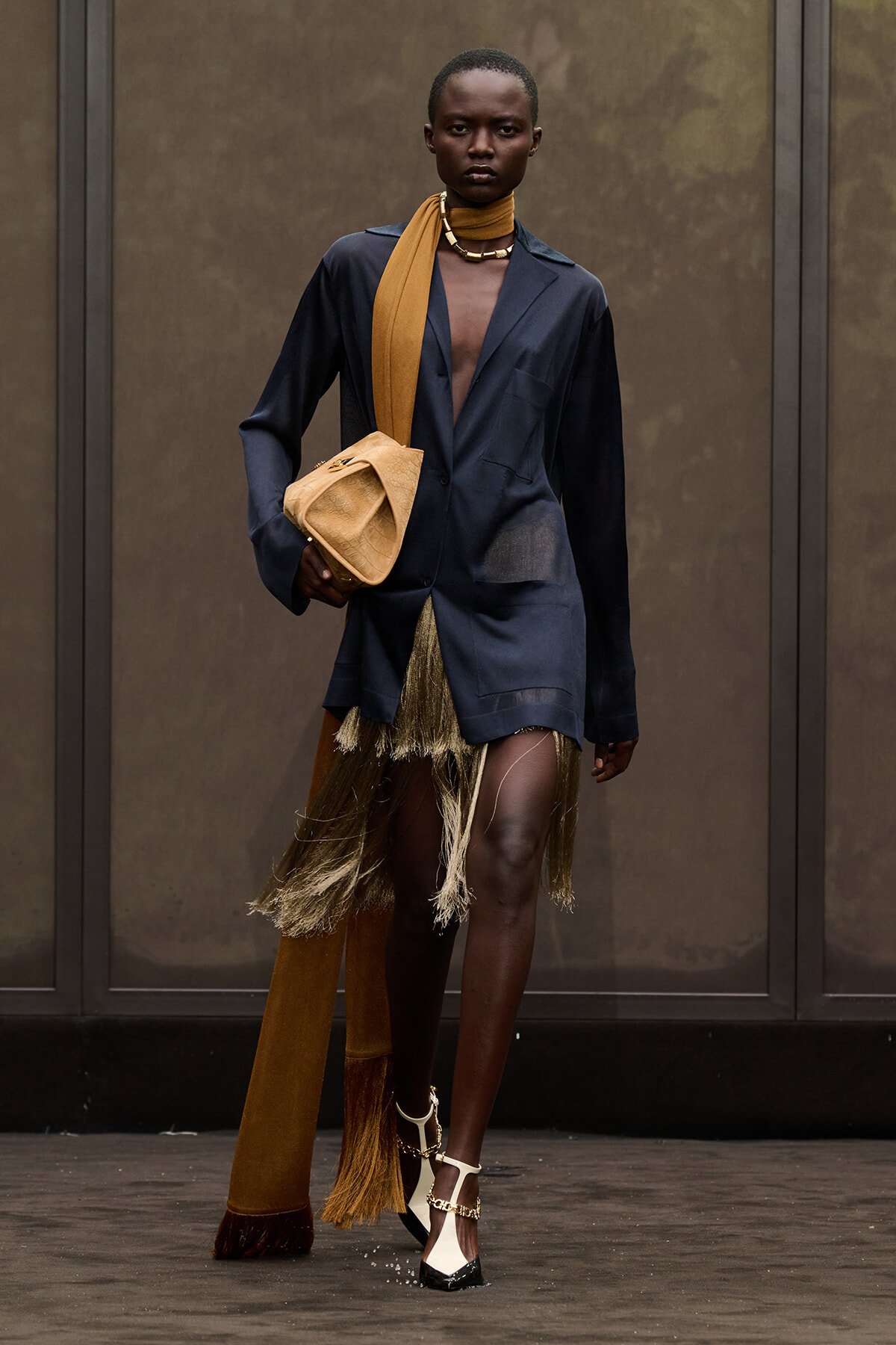 Model struts in a navy blazer-dress with gold fringe, tan scarf, and beige bag in hand.