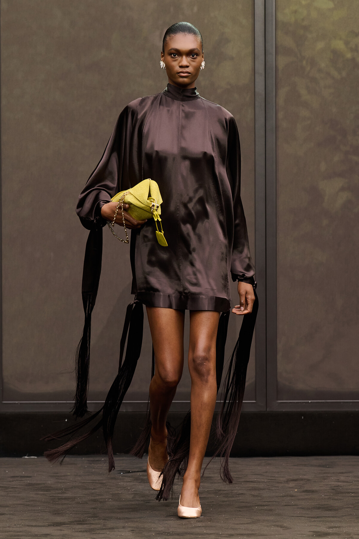 Model walking a fashion show in a glossy dark brown satin tunic with fringe accents on the sleeves and hem, holding a yellow clutch.