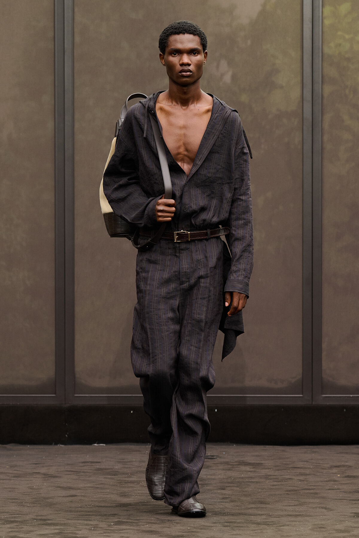 Male model walking a runway in a dark pinstripe jumpsuit, open at the chest, with a shoulder backpack.