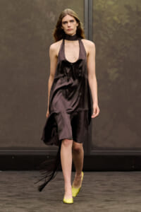 Model walking a fashion runway in a dark brown satin halter dress with a plunging neckline and asymmetric hem, paired with neon yellow flats.