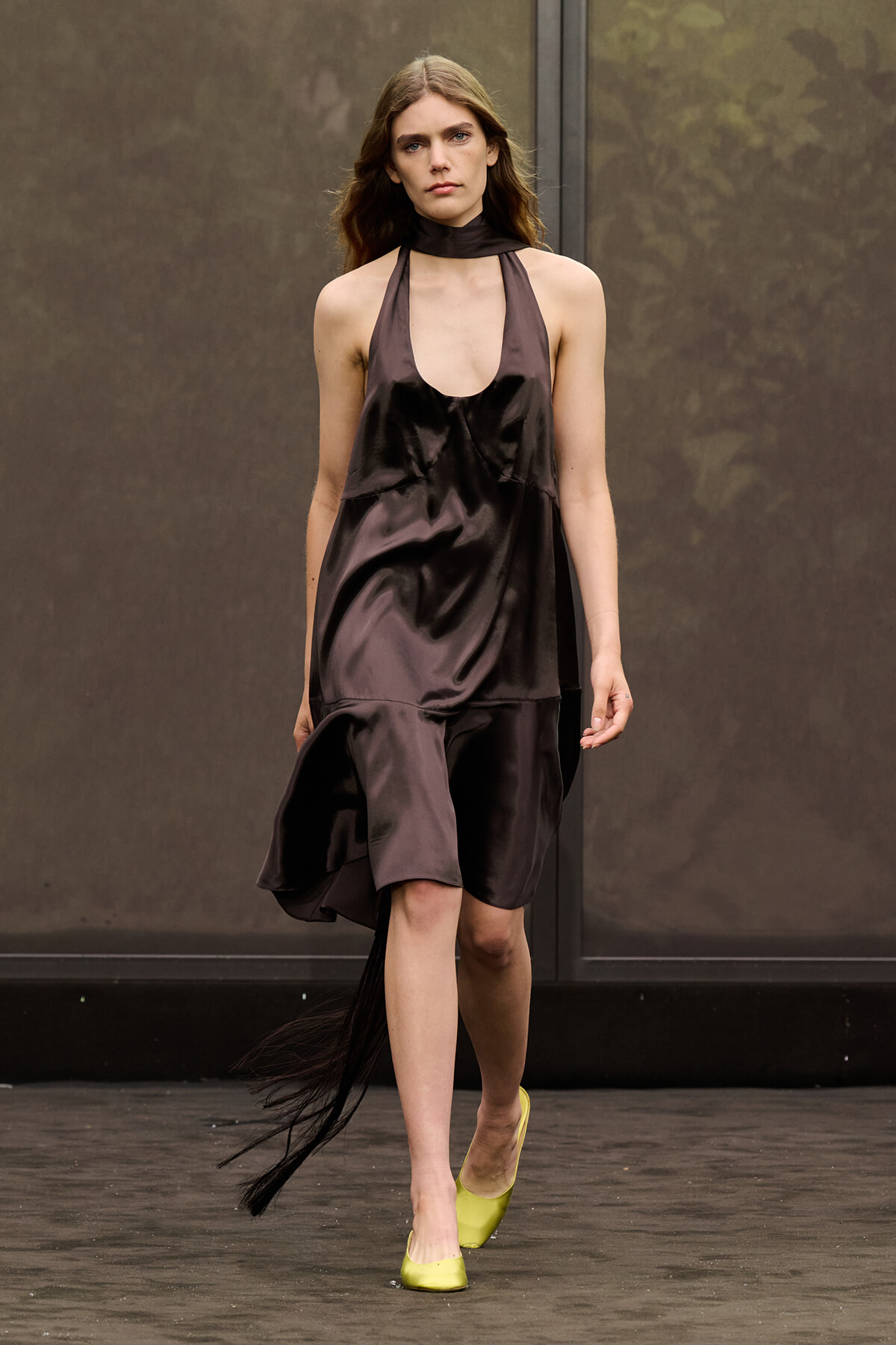 Model walking a fashion runway in a dark brown satin halter dress with a plunging neckline and asymmetric hem, paired with neon yellow flats.