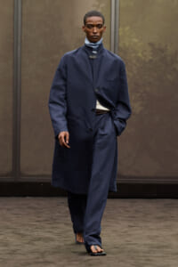 Male model walking on a runway in a long navy blue overcoat with matching trousers, belt at the waist, and sandals, serious expression against a neutral backdrop.