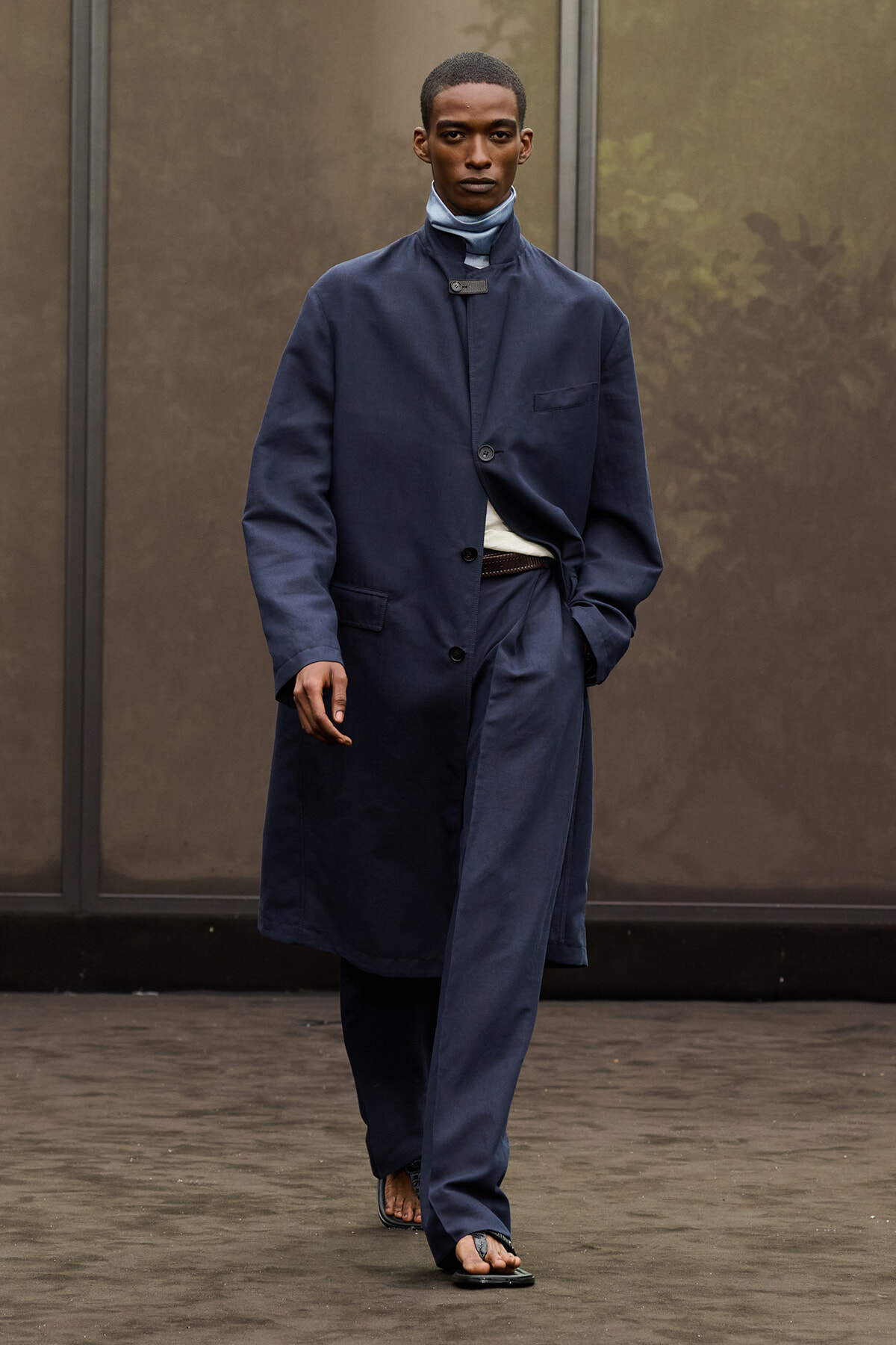 Male model walking on a runway in a long navy blue overcoat with matching trousers, belt at the waist, and sandals, serious expression against a neutral backdrop.