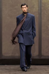 Male model in a double-breasted navy suit with a brown polka-dot scarf draped over the shoulder, walking outdoors with hands in pockets.