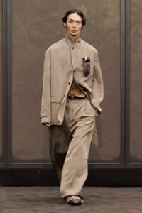 Male model wearing a loose beige suit with a brown belt and a patterned pocket square walking the runway in muted tones.