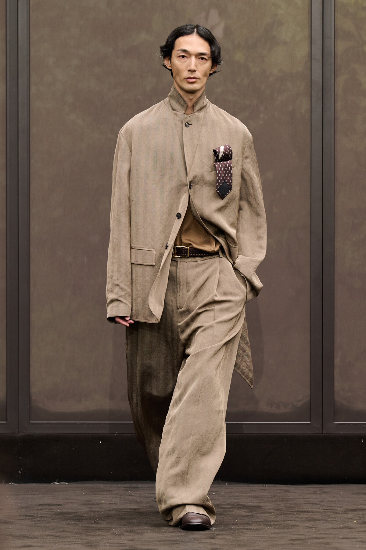 Male model wearing a loose beige suit with a brown belt and a patterned pocket square walking the runway in muted tones.