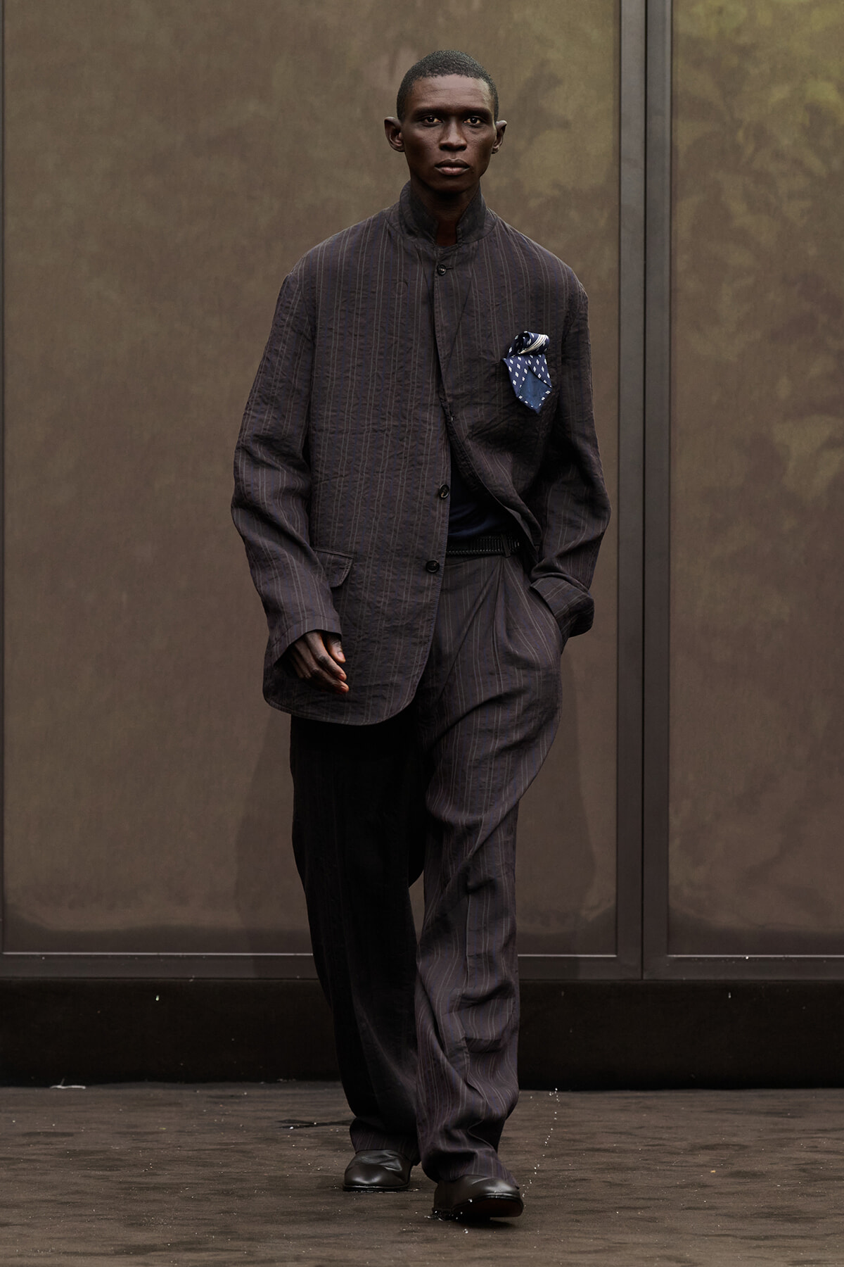 Man wearing a dark pinstripe suit with a blue pocket square, walking toward the camera.