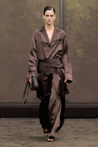 Model walking a fashion runway in a brown satin suit with a waist wrap and matching trousers, holding a small black handbag.