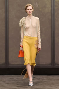 Model walking a runway in a sheer beige blouse with a gathered shoulder detail, mustard midi skirt, orange clutch, and fringe-heel shoes.