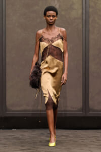 Model walking a fashion runway in a gold satin dress with brown lace panels, lime green heels, and a feathered clutch bag.