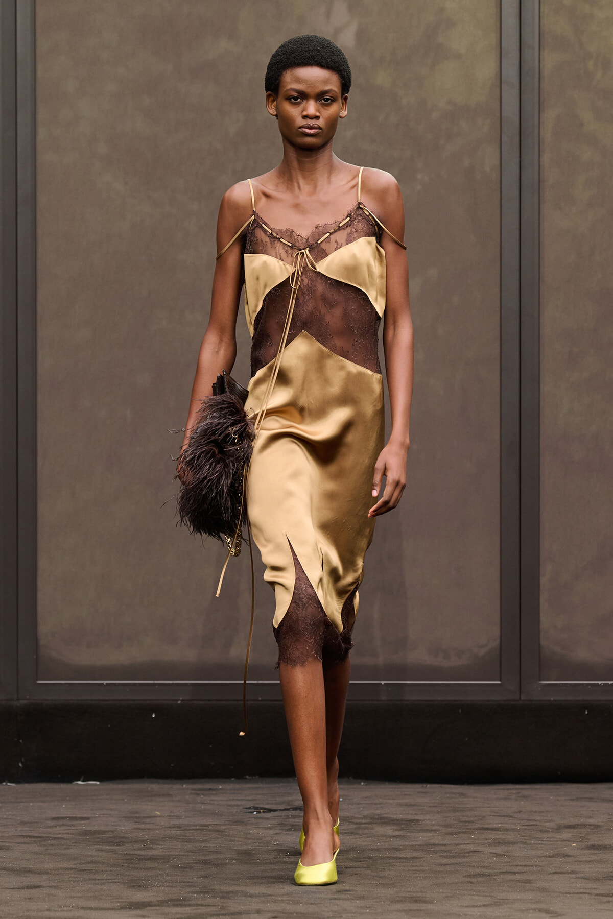 Model walking a fashion runway in a gold satin dress with brown lace panels, lime green heels, and a feathered clutch bag.
