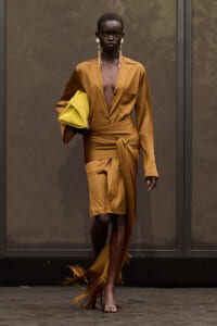Runway model wearing bronze satin blazer and wrapped skirt with a fringe hem, holding a yellow clutch.