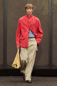 Male model wearing a red stand-collar jacket over a light blue knit, beige cargo pants, and brown shoes, carrying a yellow handbag.