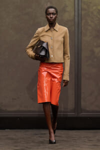 Model walking a runway in a tan cropped jacket, orange patent skirt, black handbag, and black heels.
