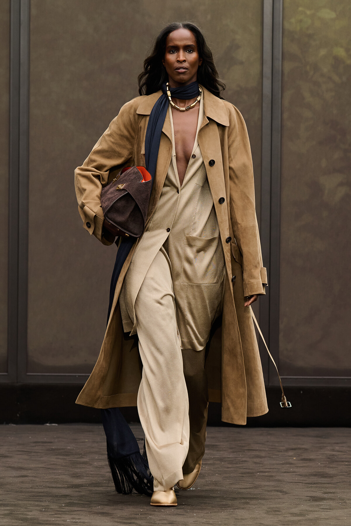 Model walking the runway in a camel trench over a beige suit, with a dark scarf and gold jewelry, carrying a brown suede bag.