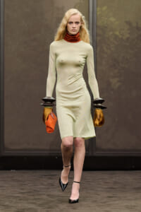 Model on a runway wearing a pale beige long-sleeve dress with a burnt-orange turtleneck, oversized black disc-style wrist cuffs and fur tassels, black heels.