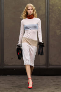 Model walking a runway in a pale pink color-block midi dress with a blue chest band and beige waist, red heels, and black circular accessories on her wrists.