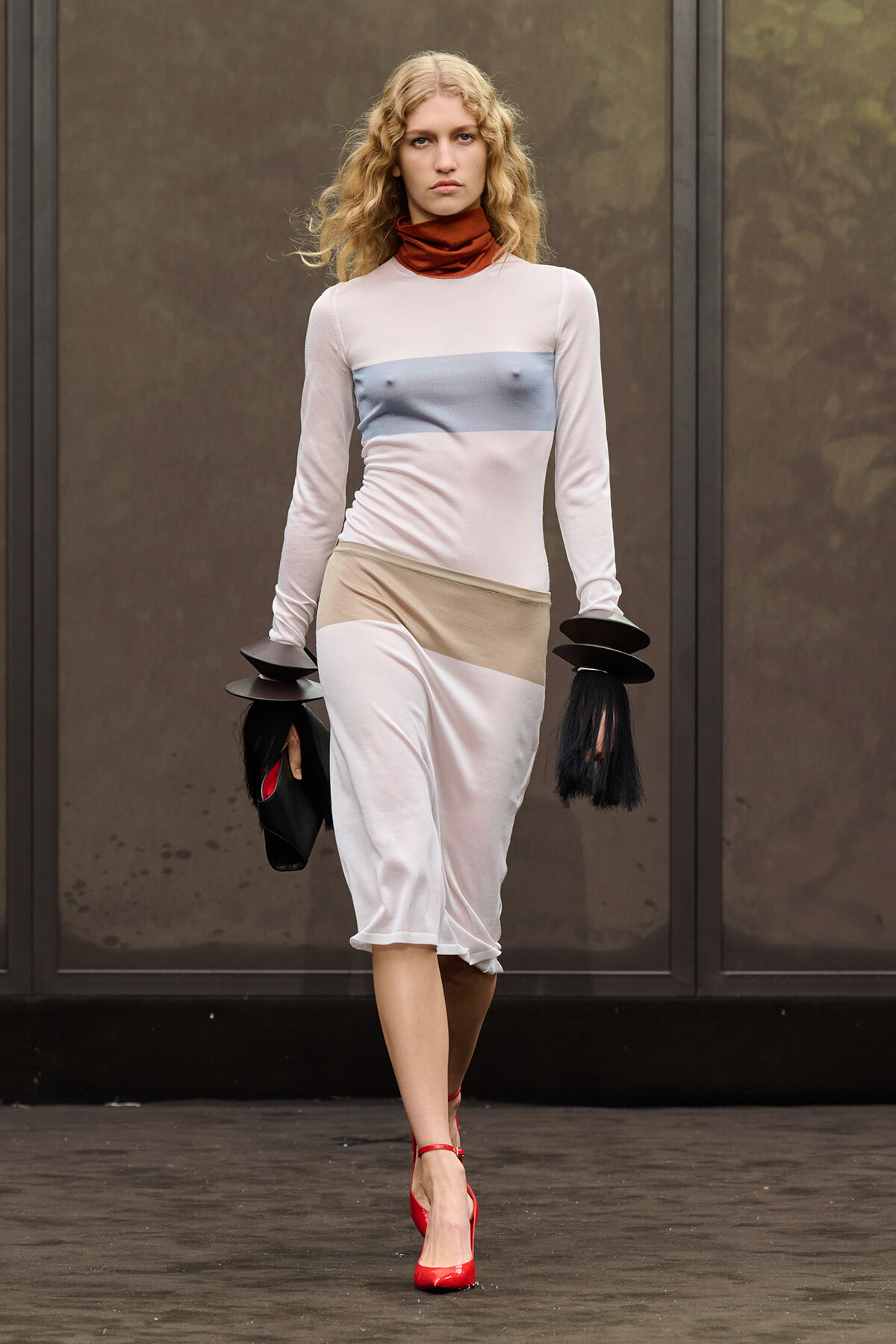 Model walking a runway in a pale pink color-block midi dress with a blue chest band and beige waist, red heels, and black circular accessories on her wrists.