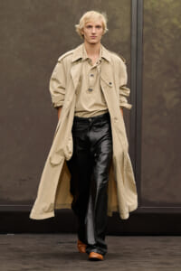 Male model walking a runway in a beige trench coat over a tan top and black leather pants, brown shoes; neutral backdrop.