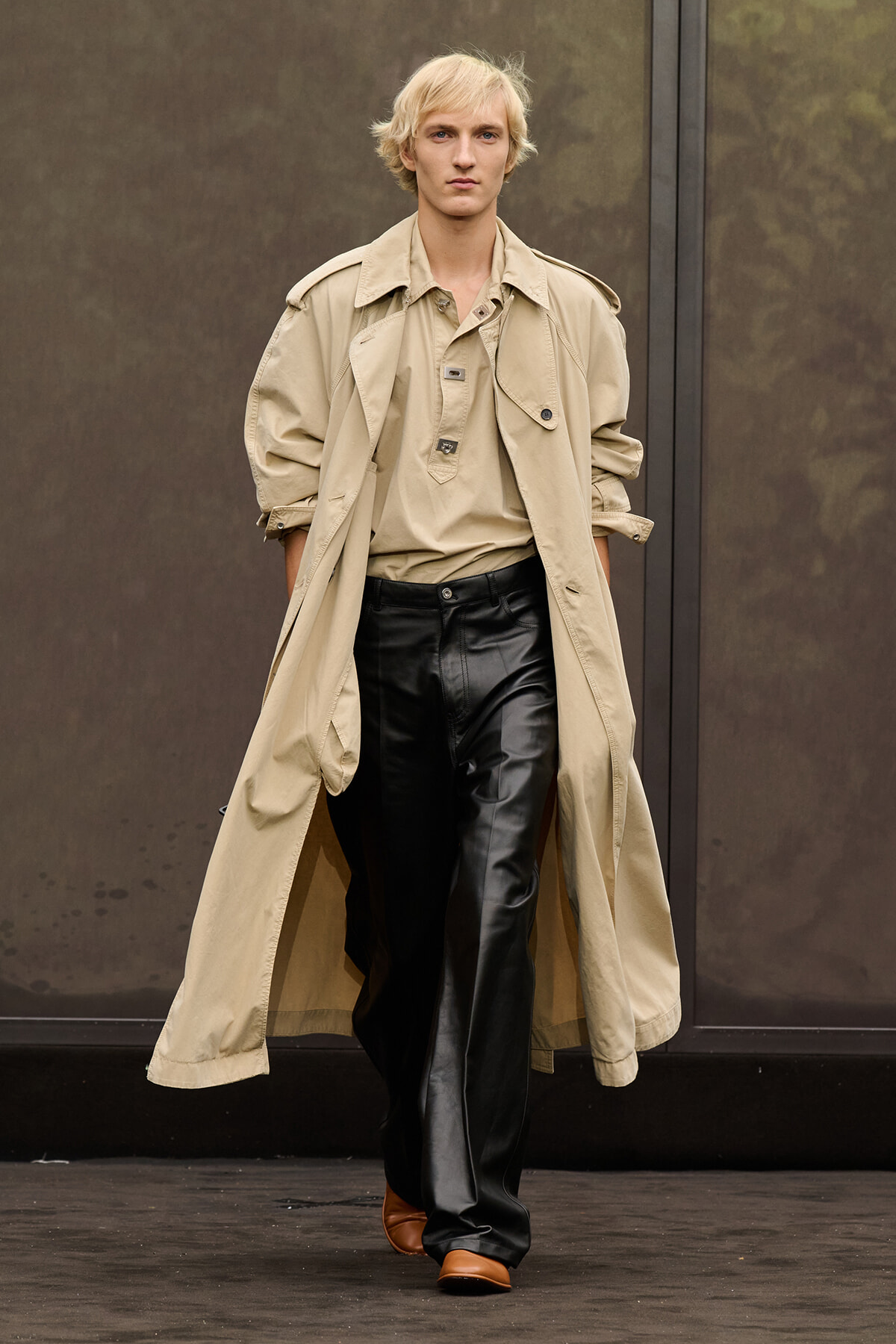 Male model walking a runway in a beige trench coat over a tan top and black leather pants, brown shoes; neutral backdrop.