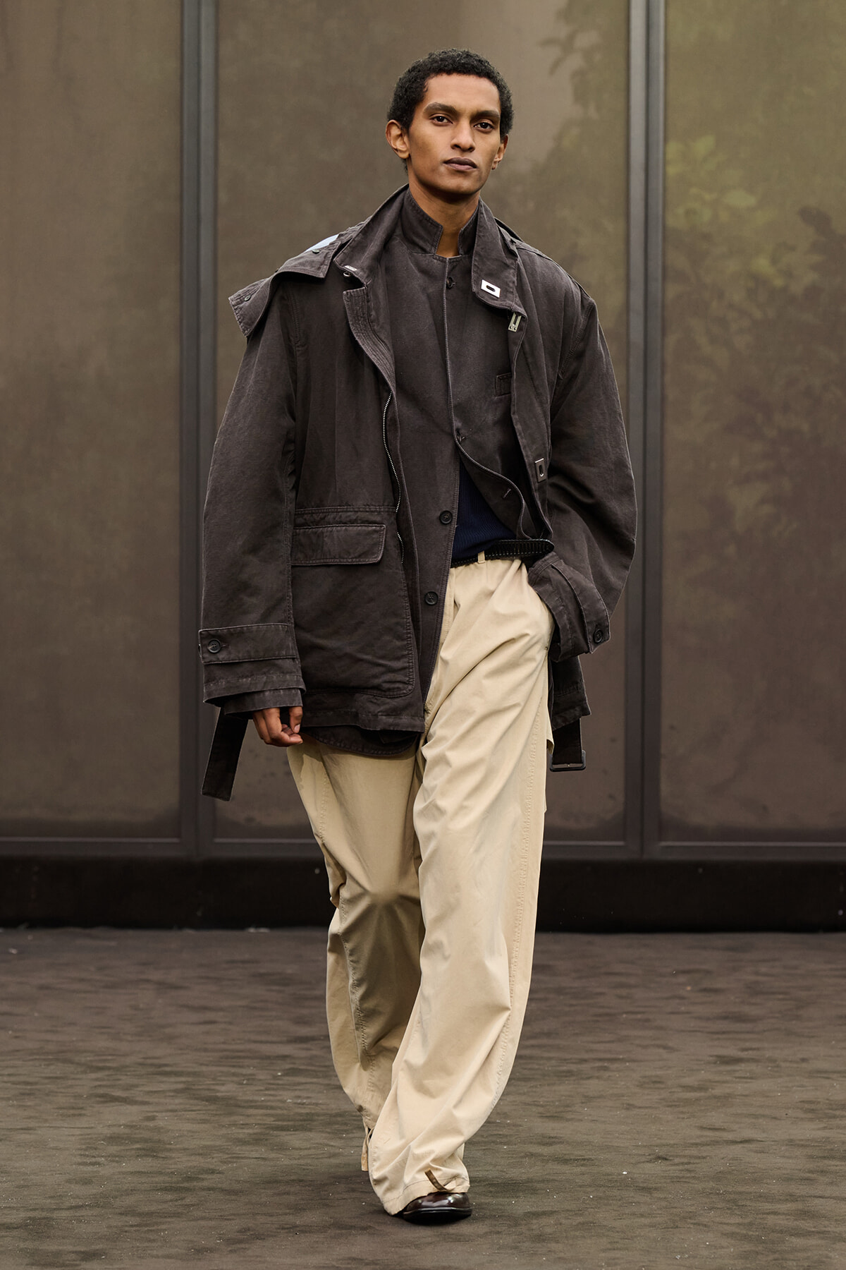 A person walking outdoors in a loose dark jacket and beige wide-leg pants, looking ahead.
