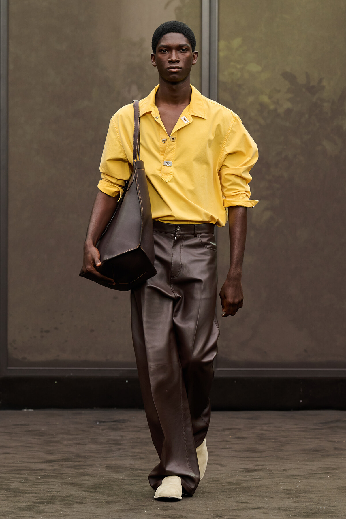 Male model walking outdoors in a yellow button-down shirt, brown leather trousers, and cream shoes, carrying a large brown shoulder bag.