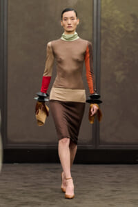 Model walks the runway in a color-block beige-brown dress with contrasting red and orange sleeves, pale green turtleneck, and tan heels, holding a beige clutch with black stacked wrist accessories.