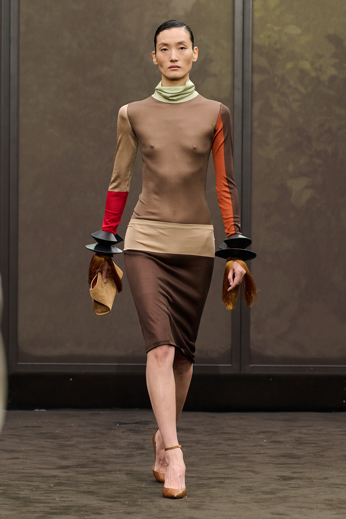Model walks the runway in a color-block beige-brown dress with contrasting red and orange sleeves, pale green turtleneck, and tan heels, holding a beige clutch with black stacked wrist accessories.